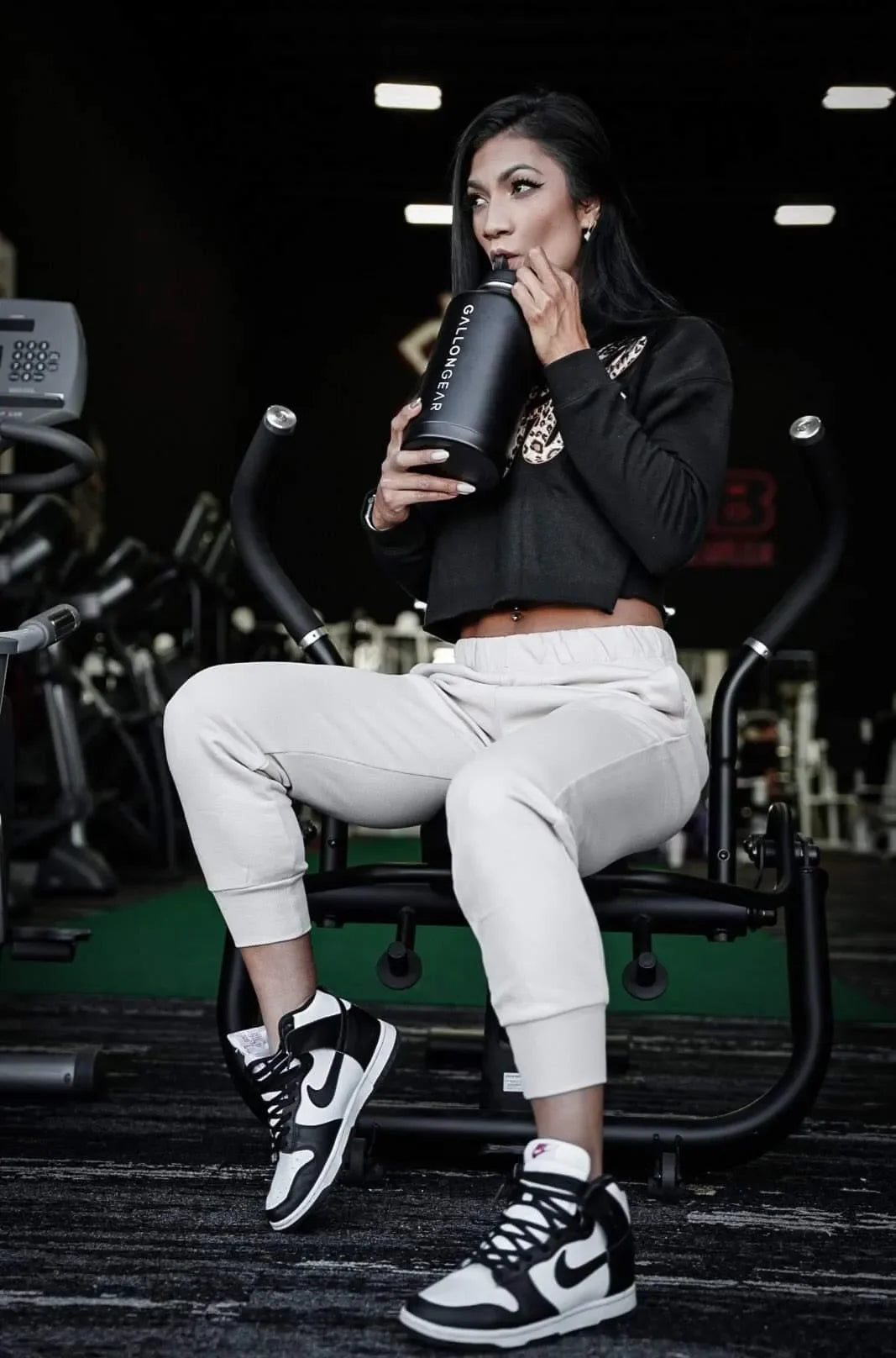 Woman drinking from Gallon Gear gym water bottle during workout, wearing athletic wear and sneakers