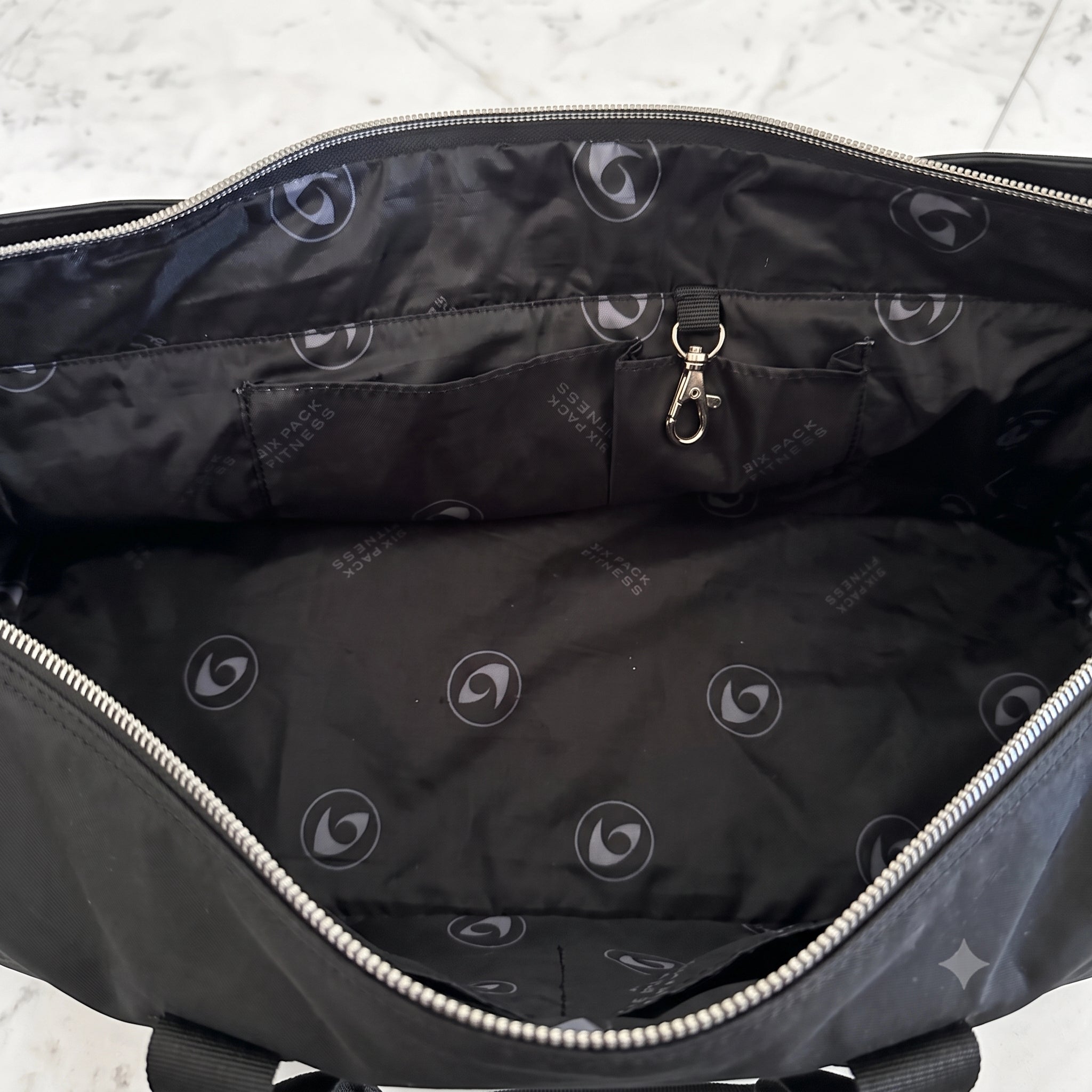 The interior of a black woman's bag shown from above, featuring a black nylon lining with a repeating light gray circular logo pattern, silver metal zippers, and a small zippered pocket with a silver key ring.