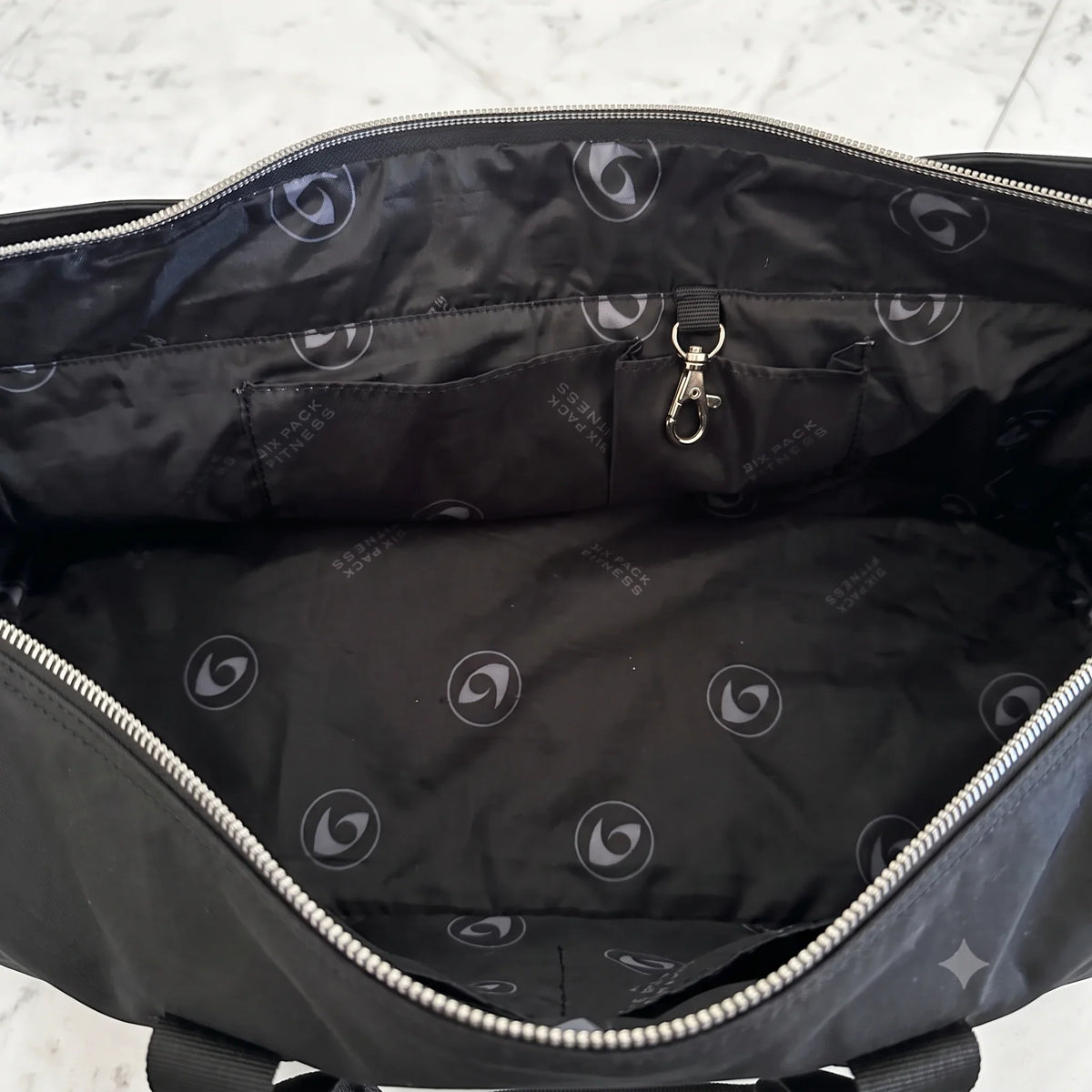 The interior of a black woman's bag shown from above, featuring a black nylon lining with a repeating light gray circular logo pattern, silver metal zippers, and a small zippered pocket with a silver key ring.