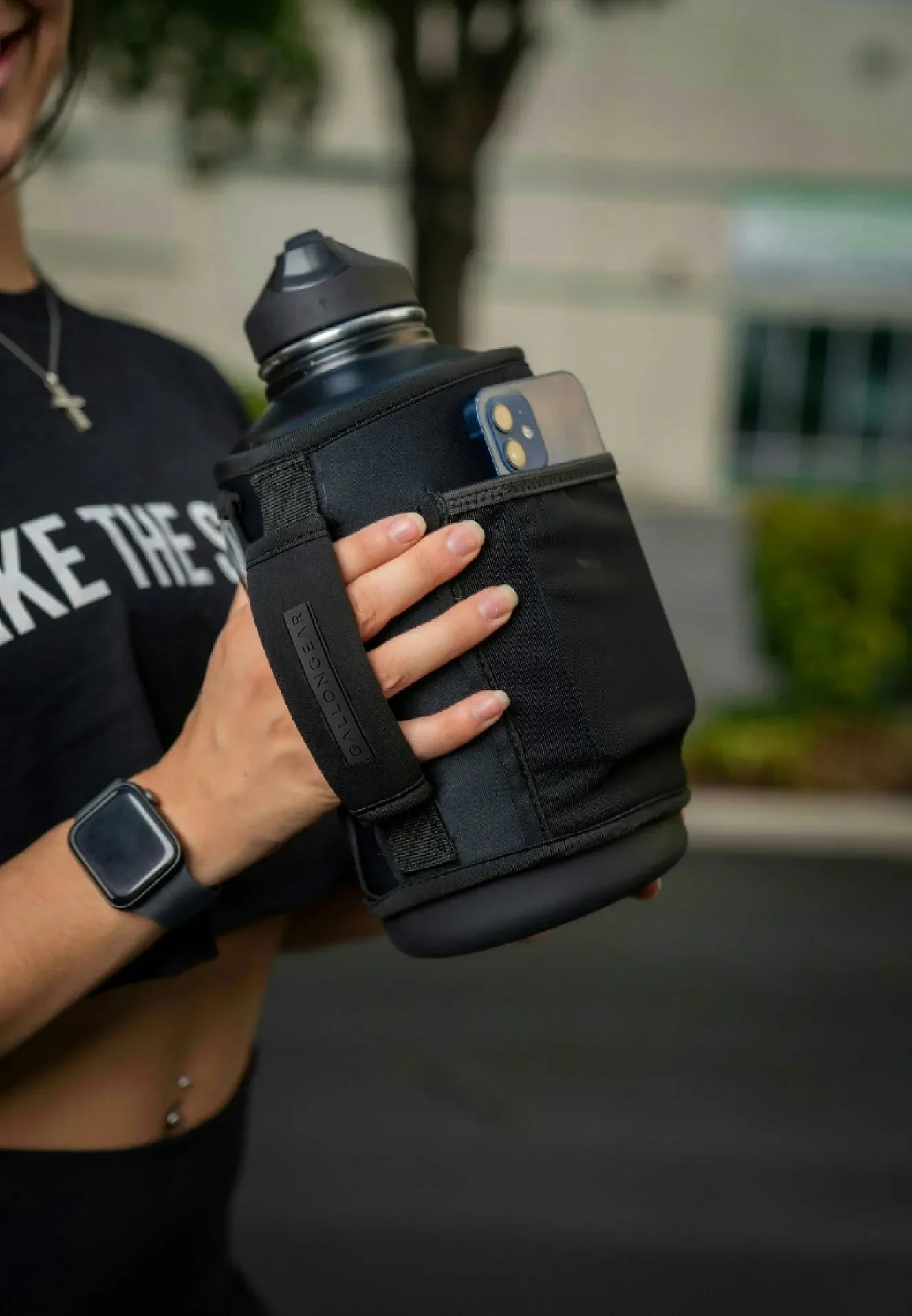 Black gallon water bottle with phone pocket, held by woman in fitness gear for gym or outdoor use